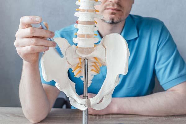 Cropped image shows a model of the spine and hip bones; a doctor uses a pen to point to the sacroiliac joint.