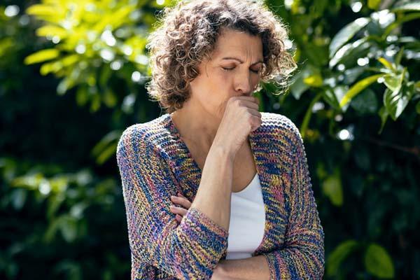 A middle-age woman in a garden covers her mouth as she coughs.