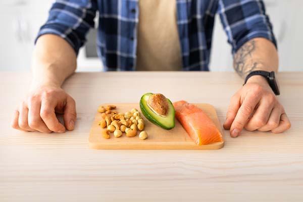 Cropped image shows the torso of a man standing next to a cutting board with avocado, assorted nuts, and salmon—all sources of healthy fats.