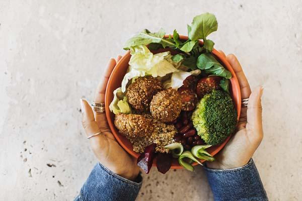 Overhead view of a woman's hands holding a bowl containing healthy, gluten-free foods: falafel, broccoli, greens, cherry tomatoes, red beans, beetroot, and quinoa.