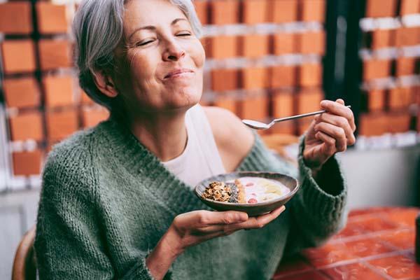 A woman smiles as she holds a bowl of cereal in one hand and a spoon in the other.