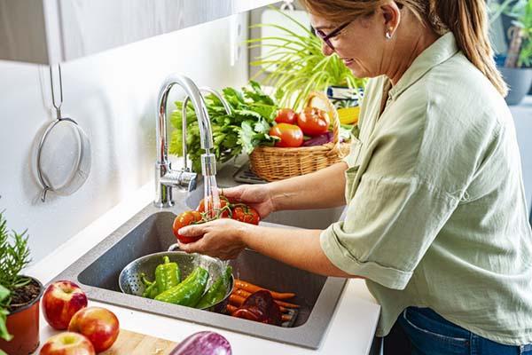 A woman washes vegetables at a sink under running water: she holds a cluster of tomatoes, there are peppers in a colander, and carrots on the bottom of the sink.