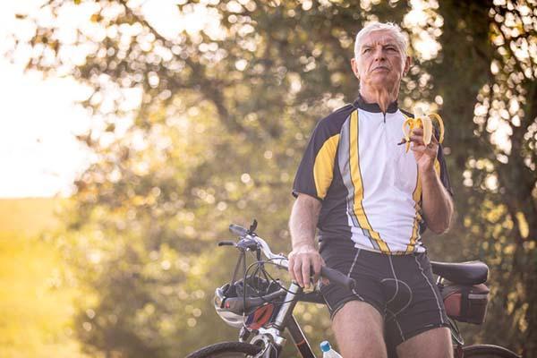 A senior man takes a break from cycling to eat a banana while leaning against his bicycle.