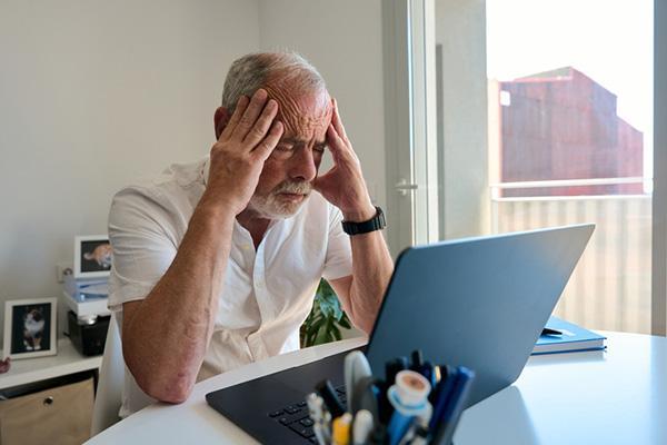 An older man working on a laptop at his home office desk holds his hands to the sides of his head, feeling a headache coming on.