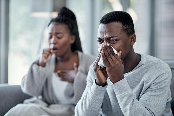 A couple at home dealing with illness: the man blows his nose into a tissue while the woman coughs and holds a hand in front of her mouth.