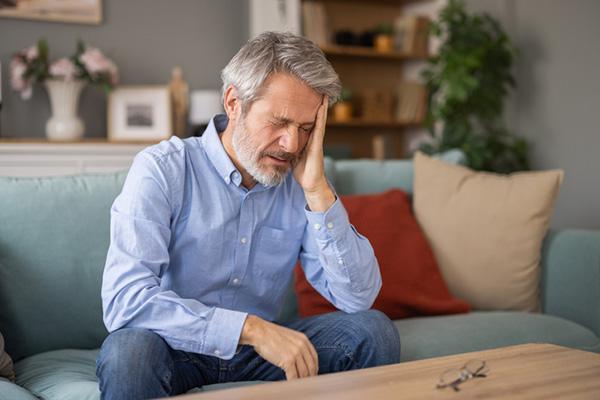 A man suffering from a severe headache holds one hand to his head while sitting on a sofa at home.