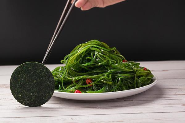 A hand holds tweezers with a disc of compressed kelp, next to a plate of kelp with grape tomatoes.