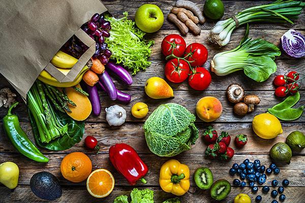 A brown paper shopping bag spilling out assorted fruits and vegetables onto a wood surface, viewed from overhead.