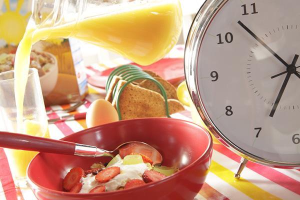 A bowl of fruit and yogurt, orange juice being poured into a glass, and a rack of toast slices all next to a large alarm clock reading five minutes to seven.