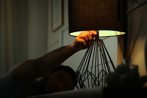 A man in bed reaches up to turn off a lamp on a bedside table.