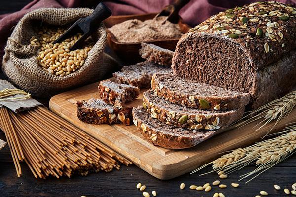 Close-up of a loaf of whole-grain bread with several slices cut, on a cutting board surrounded by other whole-grain foods.