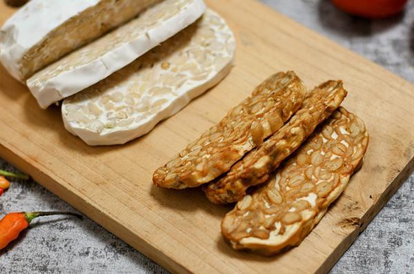 Close-up of slices of raw and fried tempeh on a wood cutting board.