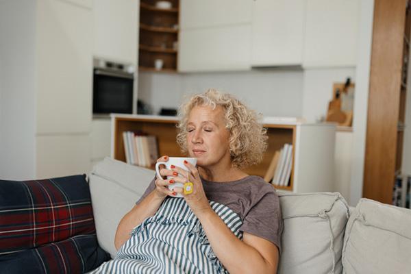 A woman sits on the sofa in her home, holding a mug and covered with a blanket.