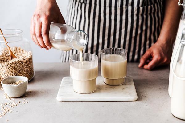 Hands of a person pouring oat milk from a bottle into a glass, with a large glass canister of oats to the side with a wood spoon in it.