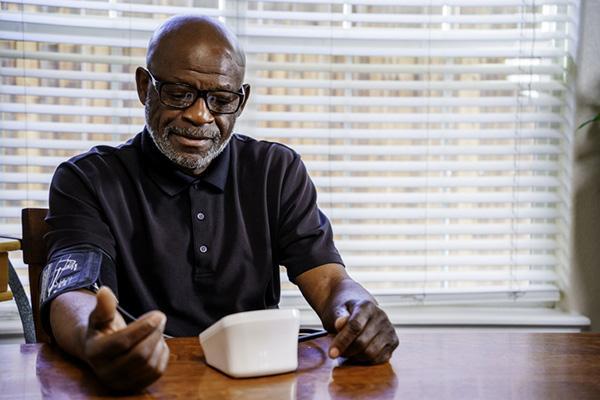 An older man sits at a table, checking his blood pressure with an arm cuff attached to an electronic monitor.