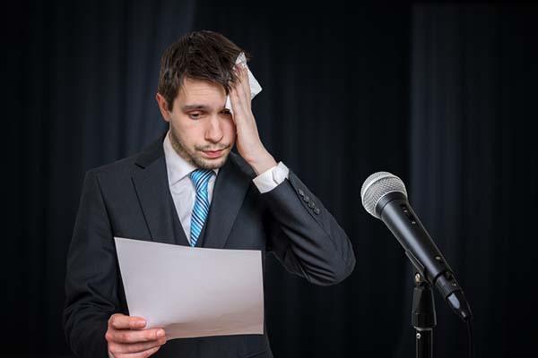 A nervous-looking man stands at a microphone holding a sheet of paper, and using a handkerchief to wipe sweat from his forehead.