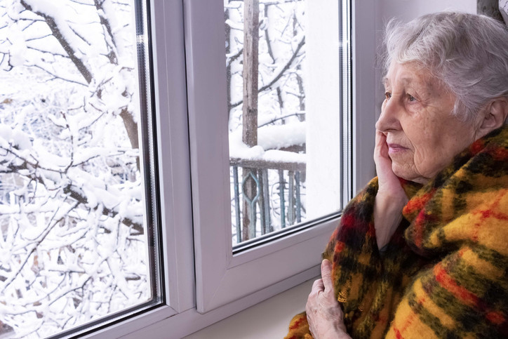 An elderly woman looking at the winter weather outside of her window.